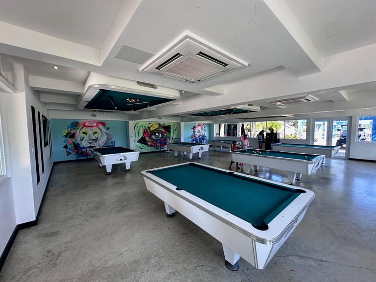 Modern recreation room with white and teal pool tables, colorful murals, and mirror-lined walls under geometric ceiling lighting