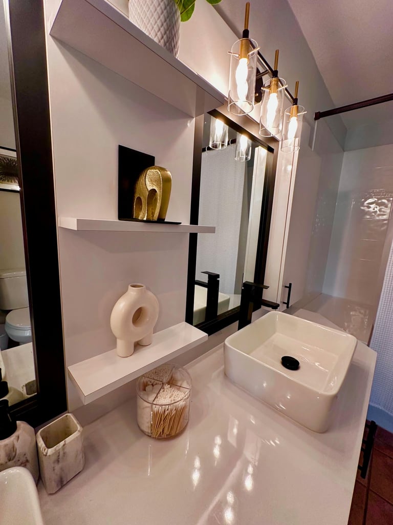 Modern bathroom with white marble vanity, white rectangular sink, brass and black pendant light fixture, and shelves displaying decorative gold and ceramic items