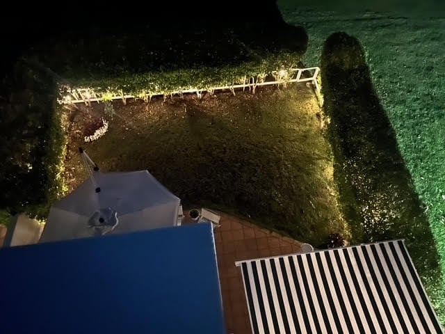 Aerial view of a backyard at night with blue pool, striped deck chair, illuminated hedges, and manicured lawn