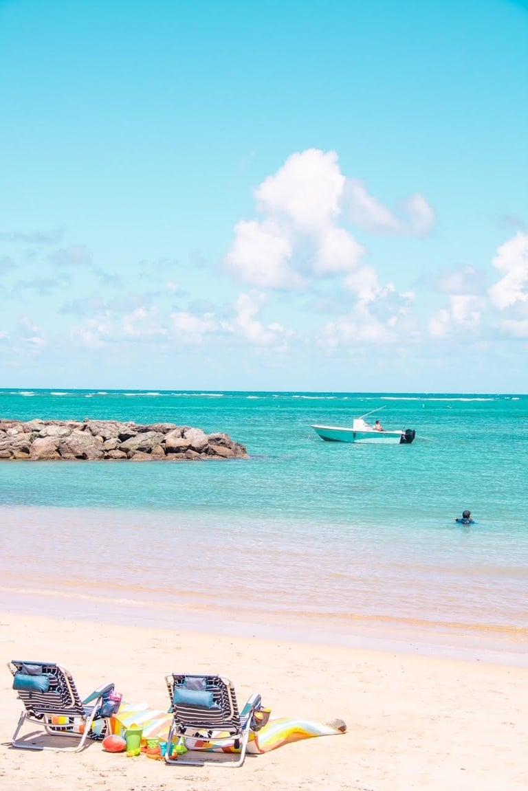 Tropical beach scene with turquoise water, sandy shore, beach loungers, a small boat, and white clouds in the sky