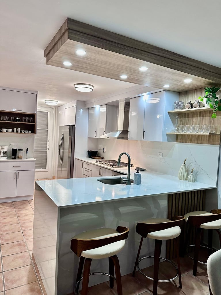 Modern white kitchen with island featuring a waterfall edge countertop, bar seating, recessed lighting, and stainless steel appliances