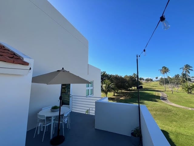 Modern white rooftop patio with gray umbrella and white seating overlooking green fields and palm trees under clear blue sky