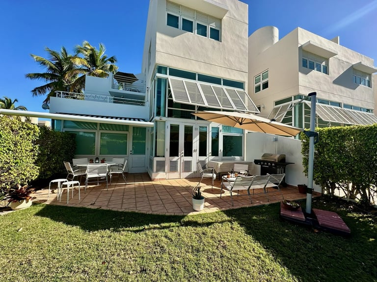 Modern beige residential home with green-framed patio doors, brick terrace, lawn, and palm trees under blue sky