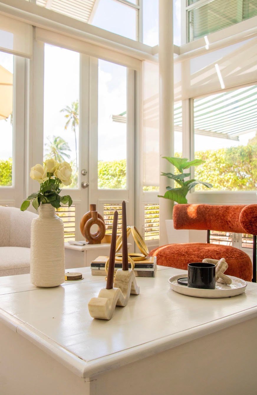 Bright sunroom with white table, potted plants, decorative objects, and rust-colored accent chair near large windows with shutters overlooking garden