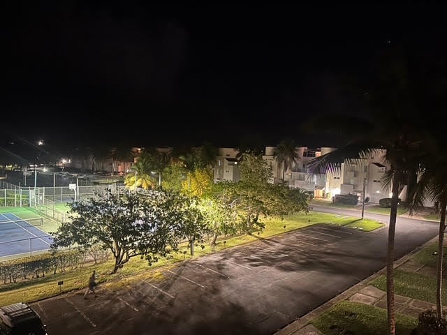 Night view of illuminated outdoor sports court with trees, pathways, and residential buildings in the background