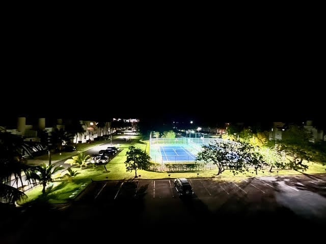 Aerial nighttime view of an illuminated tennis court surrounded by palm trees, parking lot, and buildings
