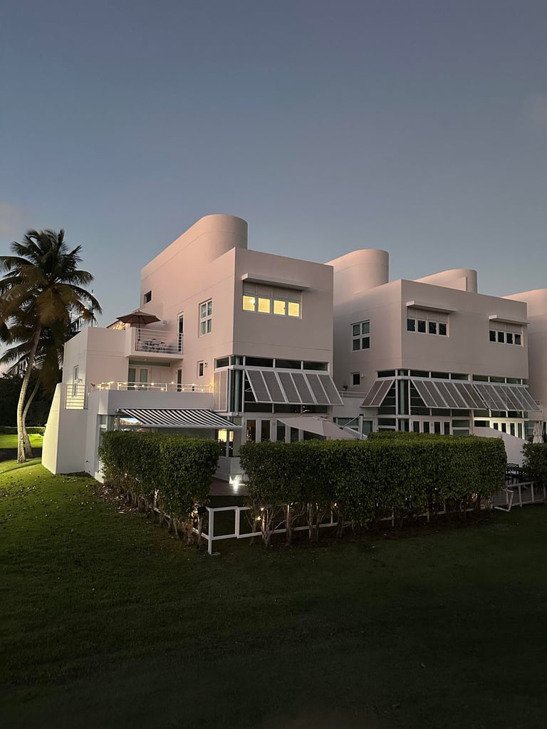 Modern white waterfront townhouses at dusk with illuminated windows, palm tree, manicured lawn, and metal railings