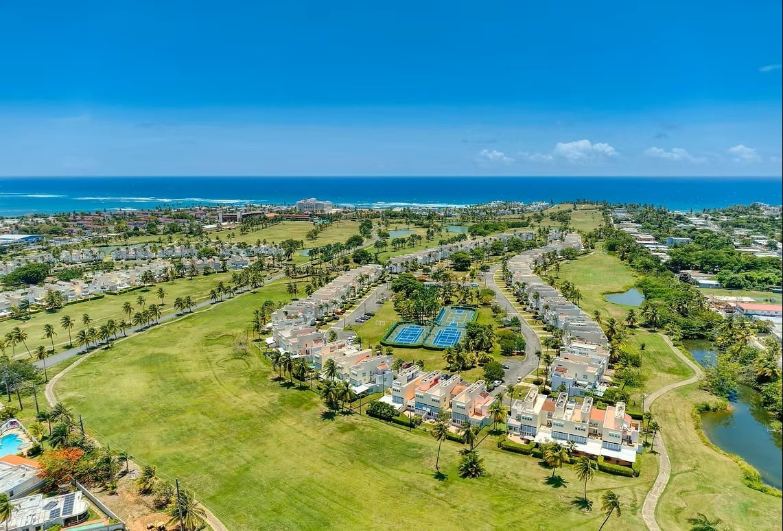 Aerial view of a coastal residential resort community with green lawns, swimming pools, and buildings overlooking the ocean