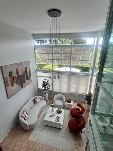 Modern living room with curved white sofa, red sculptural chairs, floor-to-ceiling windows, and hanging linear chandelier