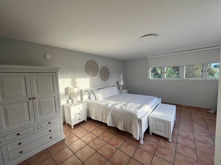Bright minimalist bedroom with white bed, wooden wardrobe, and terracotta tile floor, decorated with woven wall hangings