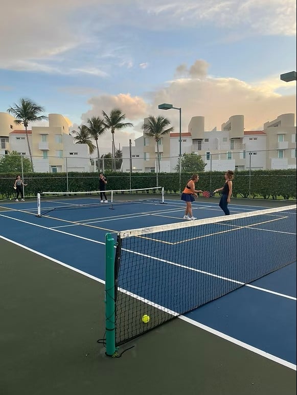 People playing tennis on blue and green courts with palm trees and white apartment buildings in the background during sunset