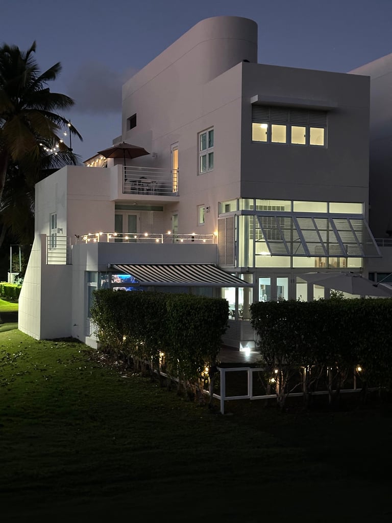 Modern multi-story house with white and dark gray exterior, lit windows, and landscaped grounds at dusk