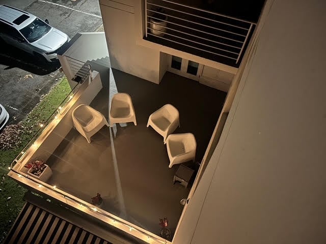 Overhead view of small room with four light-colored chairs on dark floor, visible window with air conditioning unit, and parked white car outside